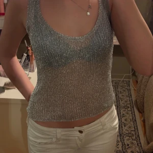 Glittrig silver linnetopp - Säljer en glittrig silverfärgad linnetopp med ribbad struktur. Toppen är ärmlös och har en rundad urringning. Perfekt för dig som vill sticka ut med en snygg och trendig look.