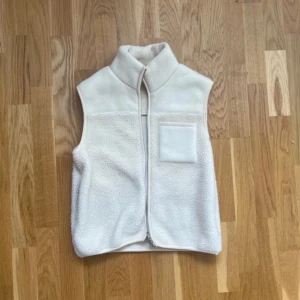 Vit Teddy väst - Mysig vit fleeceväst med hög krage och dragkedja framtill. Västen har en enkel design med en liten bröstficka på vänster sida och är tillverkad i mjukt, fluffigt fleecematerial. Perfekt att slänga på sig över en hoodie eller långärmad tröja.