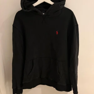 Ralph Lauren hoodie  - Svart hoodie från Polo Ralph Lauren, Klassisk design med huva, känguruficka och röd broderad logga på bröstet. Tillverkad i mjuk bomull, perfekt för chill dagar. Snygg och enkel att matcha med allt. Storlek XL men passar L