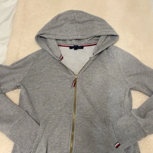 Grå hoodie från Tommy Hilfiger - Snygg grå hoodie från Tommy Hilfiger med dragkedja i guld och klassiska Hilfiger-detaljer vid dragkedjan och ärmslut. Tröjan har huva, långa ärmar och två fickor framtill. Perfekt för en chill och sportig look.