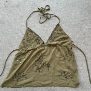 Beige linnetopp med pärlor och paljetter - Säljer en beige linnetopp med tunna knytband i nacken och v-ringad front. Toppen har broderade detaljer med pärlor och paljetter i silver som bildar ett mönster över hela plagget. Materialet är tunt och luftigt, perfekt för varma dagar.Liite trasig på kanten men syns ej