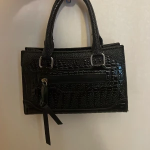 Svart handväska i croco-mönstrat skinn - Snygg svart handväska med croco-mönstrad yta och silvriga metalldetaljer. Väskan har två handtag, dragkedja upptill och ett extra fack med dragkedja på framsidan. Perfekt storlek för det viktigaste och ger en cool vibe till din outfit.
