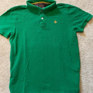 Grön pikétröja från Morris, M - Snygg grön pikétröja från Morris med klassisk krage och två knappar framtill. Tröjan har en broderad guldfärgad logga på bröstet och är gjord i 100% bomull. Perfekt för en stilren och avslappnad look.