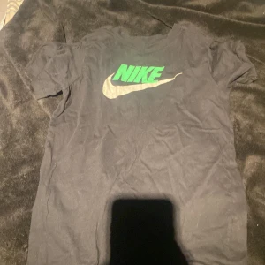 Svart Nike t-shirt med grön logga - Svart t-shirt från Nike med klassisk grön Nike-logga och silverfärgad swoosh på bröstet. T-shirten är i bomull och har en normal passform med korta ärmar. Perfekt för dig som gillar sportiga och stilrena plagg.