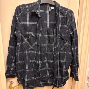 Svart rutig skjorta från H&M Stl 42 - Svart långärmad skjorta från H&M Divided i storlek 42. Skjortan har klassisk krage, två bröstfickor med lock och knappar framtill. Tillverkad i mjuk bomull, perfekt för en avslappnad och cool look.