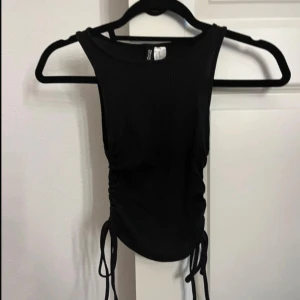 Svart ribbad linnetopp med snörning - Svart ärmlös ribbad linnetopp från Gina Tricot med snörning i sidorna som ger en snygg croppad look. Toppen har rund halsringning och är perfekt för dig som gillar stilrena basplagg med en twist.