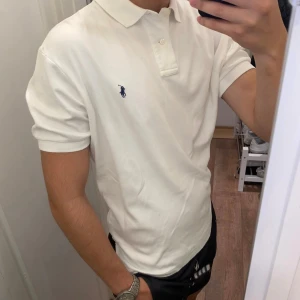 Vit pikétröja från Polo Ralph Lauren - Vit pikétröja från Polo Ralph Lauren i 100% bomull. Klassisk design med krage, två knappar och korta ärmar. Ikoniska blå logon broderad på bröstet. Perfekt för en clean och stilren look, passar dig som gillar tidlös stil. Small men passar Märkt Small men sitter som en lätt Medium.
