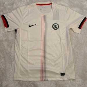 Chelsea FC vit Nike fotbollströja - Chelsea FC fotbollströja från Nike i vit polyester med tunna vertikala ränder i svart och rött på framsidan. Kortärmad modell med svarta och röda detaljer på ärmsluten samt klubbmärke på bröstet. Snygg och sportig design för fans.