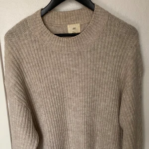 Beige ribbstickad tröja från H&M - Mysig beige ribbstickad tröja från H&M i storlek M. Tröjan har en rund halsringning och långa ärmar, perfekt för lager på lager under kyliga dagar. Enkel och stilren design som funkar till det mesta.