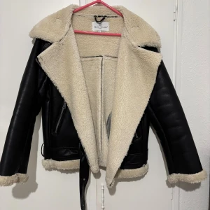 Svart teddyjacka från Melva Melody - Snygg svart teddyjacka från Melva Melody med krämvit fuskpäls på insidan och krage. Jackan har dragkedjor på fickorna, bälte i midjan och långa ärmar med pälsdetaljer vid ärmslut och nederkant. Perfekt för kalla dagar och ger en cool look.