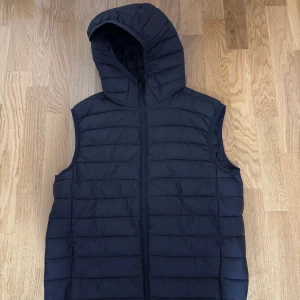 Svart dunväst med huva - Snygg svart dunväst med huva och horisontella quiltade sömmar. Västen har dragkedja framtill och är perfekt för lager-på-lager. Enkel, stilren design som funkar till många olika outfits. Passar dig som gillar en sportig och chill look.