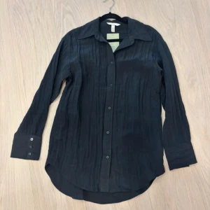 Svart skjorta från H&M  - Svart skjorta från H&M med lätt skrynklig struktur. Klassisk krage, knäppning framtill och långa ärmar med manschett och knappar. Skjortan har en rak och lite längre passform, perfekt för en clean och stilren look. Etikett kvar. Aldrig använd. Storlek XS. 