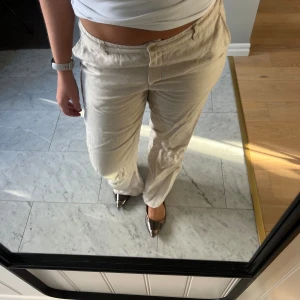 Beiga linnebyxor med rak passform - Snygga beiga linnebyxor med rak och lite loose passform. Byxorna har medel hög midja, klassiska fickor och stängs med knapp och dragkedja. Perfekta för dig som gillar en chill och stilren look. Materialet är linne som ger en skön och luftig känsla.