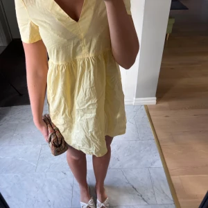 Gul kort klänning med v-ringning - Söt ljusgul kort klänning med v-ringad hals och korta ärmar. Klänningen har en avslappnad passform med rynkad midja och är tillverkad i ett lätt bomullstyg, perfekt för varma dagar. Enkel och stilren design som känns fräsch och somrig.