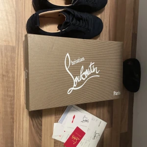 Christian Louboutin svarta sneakers - Säljer ett par svarta sneakers från Christian Louboutin i mocka med den ikoniska röda sulan. Skorna har snörning och rund tå, och kommer med originalkartong och äkthetsbevis. Perfekta för dig som vill sticka ut med exklusiva detaljer.