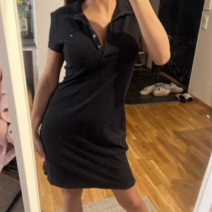 Svart kort klänning Tommy Hilfiger - Säljer en svart kort klänning från Tommy Hilfiger i slim fit-modell. Klänningen har krage, knappar framtill och korta ärmar. Klassisk och enkel design med liten logga på bröstet. Perfekt för dig som gillar stilrena plagg med sportig touch.