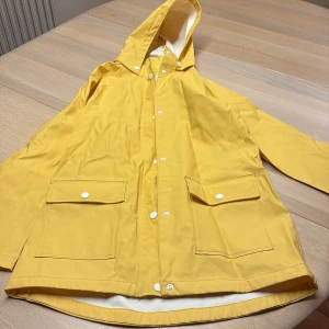 Gul regnjacka med huva från H&M - Snygg gul regnjacka från H&M med vit insida, huva och stora fickor med lock framtill. Jackan har vita knappar. Lång i modellen och fint skick 