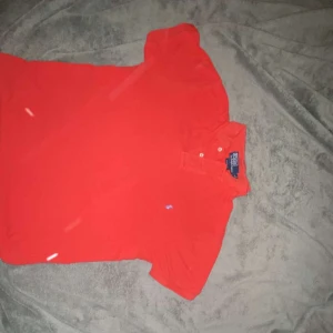 Röd pikétröja från Polo Ralph Lauren - Snygg röd pikétröja från Polo Ralph Lauren med klassisk krage och knappar framtill. Tröjan har korta ärmar och den ikoniska lilla broderade loggan på bröstet. Perfekt för en fräsch och sportig look.