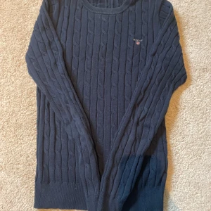 Mörkblå kabelstickad tröja från Gant - Säljer en mörkblå kabelstickad tröja från Gant med rund halsringning och diskret logga på bröstet. Tröjan är tillverkad i stretchig bomullsblandning och har långa ärmar samt ribbade muddar för en klassisk look.