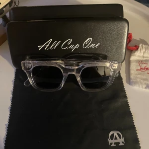 Klara solglasögon från All Cap One - Säljer ett par stilrena solglasögon från All Cap One med transparent båge och svarta glas. Rektangulär form som ger en modern vibe. Kommer med originalfodral och putsduk med logga. Perfekt accessoar för att lyfta din look.