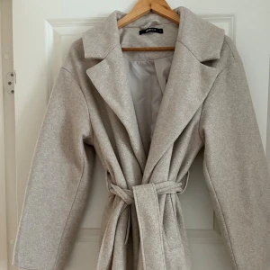 Beige kappa från Ginatricot med bälte - Snygg beige kappa från Ginatricot med klassisk krage och knytbälte i midjan. Jackan har lång ärm och en stilren, rak passform som ger en clean look. Perfekt för dig som gillar minimalistisk stil och vill ha en jacka som funkar till många outfits.