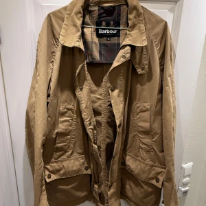 Beige Barbour parkajacka XL - Säljer en klassisk beige parkajacka från Barbour i storlek XL. Jackan har flera stora fickor med knappar, krage i manchester och rutigt innerfoder. Perfekt för dig som gillar tidlös stil och funktionella detaljer.