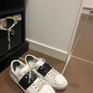 Valentino Garavani vita sneakers - Snygga vita sneakers från Valentino Garavani med bred svart läderdetalj över vristen och ikoniska nitar på hälen. Skorna har rund tå, vita snören och är tillverkade i skinn för en lyxig känsla. Perfekta för dig som vill ha en stilren och exklusiv look.