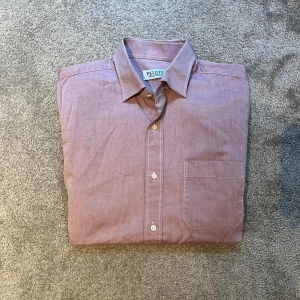 Rosa skjorta - Säljer en stilren rosa skjorta från Pelote med klassisk krage och bröstficka. Skjortan har knäppning framtill och är tillverkad i mjuk bomull. Perfekt för dig som gillar en clean och enkel look.