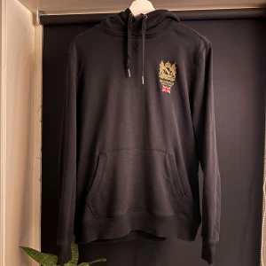 Svart hoodie från Morris Stockholm - Svart hoodie från Morris Stockholm med guldfärgat broderi och brittisk flagga på bröstet. Tröjan har huva med snörning, stor magficka och långa ärmar. Perfekt för dig som gillar stilrena och klassiska detaljer. Passa nog även S