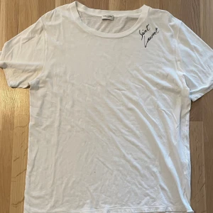 Vit Saint Laurent t-shirt i bomull - Vit t-shirt från Saint Laurent med svart logga broderad vid axeln. Klassisk rund halsringning och korta ärmar. Tillverkad i mjuk bomull för en skön och avslappnad känsla. Perfekt för dig som gillar stilrena och exklusiva plagg. Litet litet hål vid bröstet