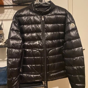 Svart Moncler pufferjacka - Säljer en svart pufferjacka från Moncler Longue Saison med klassisk quiltad design och hög krage. Jackan har dragkedja framtill, två sidofickor med dragkedja och Moncler-logga på ärmen. Materialet är glansigt och lätt, perfekt för kyliga dagar. 