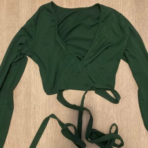 Mörkgrön omlottopp med långa ärmar - Snygg mörkgrön omlottopp med långa ärmar och knytband i midjan. Toppen har en v-ringad design och är croppad, vilket ger en trendig look. Tillverkad i mjukt bomullsmaterial som känns skönt mot huden. Perfekt att styla med höga jeans eller kjol.