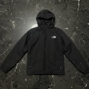 Svart vindjacka från The North Face - Svart vindjacka från The North Face med huva och logga på bröstet och ryggen. Jackan har dragkedja framtill, två sidofickor och elastiska muddar vid ärmslut. Materialet är slitstark polyester och jackan är perfekt för blåsiga dagar.