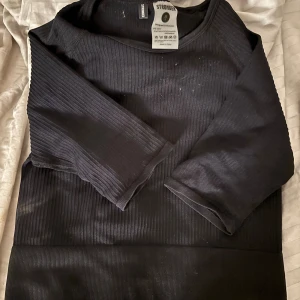 Svart ribbad träningströja Stronger XS/S - Svart trekvarts ärmad träningströja från Stronger i ribbat material med diskret logga på mudden. Tröjan är tillverkad i stretchig polyamidblandning och har en tight passform som sitter snyggt på kroppen. Perfekt för gymmet eller idrotten!🤍