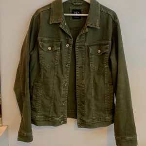 Olivgrön jeansjacka från Zara  - Snygg olivgrön jeansjacka från Zara Man i storlek L. Klassisk modell med krage, två bröstfickor med knappar och raka sömmar. Jackan är tillverkad i bomull och har silverfärgade knappar med logga. Perfekt för dig som gillar stilren och enkel design.