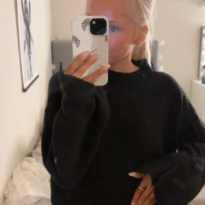 Svart stickad tröja från bpc selection - Mysig svart stickad tröja från bpc selection med oversized passform och långa ärmar. Tröjan har en enkel design med ribbade muddar och hög hals samt små volanger på ärmarna, perfekt för kalla dagar. Snygg att styla med jeans eller kjol.