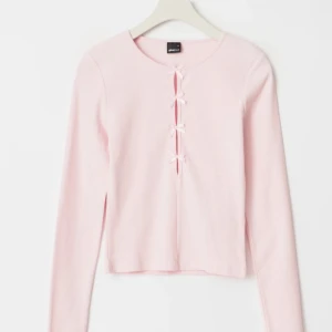 Ljusrosa ribbad topp med rosetter - Supersöt ljusrosa ribbad topp från Gina Tricot med långa ärmar och croppad passform. Framsidan har fyra små rosetter längs en öppning som ger en unik och gullig detalj. Perfekt att styla med jeans eller kjol för en trendig look. Helt ny med prislapp 