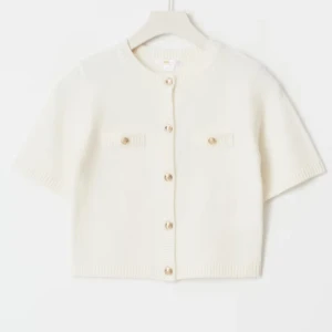 Vit kortärmad kofta med knappar - Säljer en stilren vit kofta med korta ärmar och snygga knappar framtill. Koftan har två dekorativa fickor på bröstet och är stickad i ett mjukt material. Perfekt för lager-på-lager och enkel att matcha med olika outfits. 