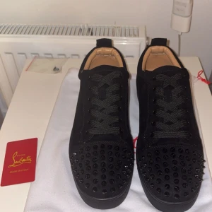 Christian Louboutin svarta sneakers med nitar - Svarta sneakers från Christian Louboutin i mocka med coola svarta nitar på tån och klassisk röd sula. Skorna har snörning och rund tå, samt lyxiga detaljer som beige innersula och medföljande dustbag. Perfekta för dig som vill sticka ut med exklusiv streetstyle.
