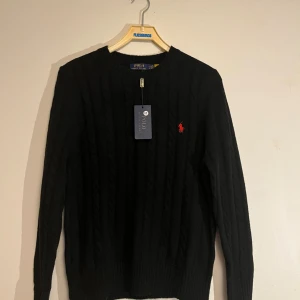 Svart kabelstickad tröja Polo Ralph Lauren - Svart kabelstickad tröja från Polo Ralph Lauren med klassisk rund hals och röd broderad logga på bröstet. Tröjan har lång ärm och ribbade muddar. Perfekt för dig som gillar stilren och tidlös design.