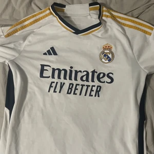 Real Madrid vit fotbollströja Adidas M - Säljer en officiell Real Madrid fotbollströja från Adidas i storlek M. Tröjan är vit med mörkblå detaljer och guldiga ränder på axlarna. Den har korta ärmar, klubbmärke på bröstet och Emirates Fly Better-tryck. Materialet är lätt och andas, perfekt för match eller träning.