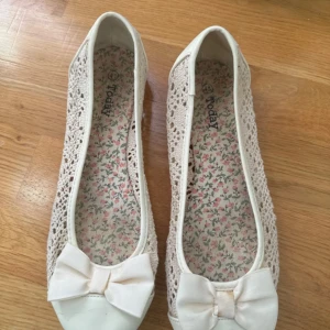 Vita ballerinaskor med rosett - Söta vita ballerinaskor från Today med spetsdetaljer och stor rosett framtill. Skorna har rund tå och insida med blommigt mönster. Perfekta för vår och sommar när du vill ha en romantisk och feminin look.