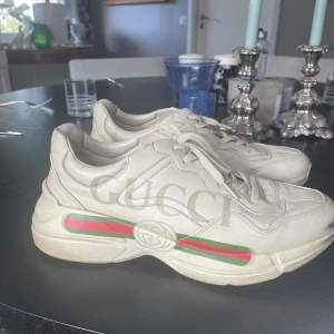 Gucci Rhyton sneakers i beige skinn - Chunky Gucci Rhyton sneakers i beige skinn med stora logotyptryck på sidan och klassiska gröna och röda ränder. Skorna har snörning, rund tå och platt sula. Perfekta för dig som vill sticka ut med en lyxig streetstyle-look.