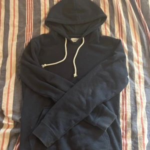 Mörkblå hoodie från Pier One - Mysig mörkblå hoodie från Pier One med klassisk huva och vita snören. Tröjan har en känguruficka framtill och långa ärmar. Perfekt för chill dagar eller när du vill ha en enkel och stilren look.