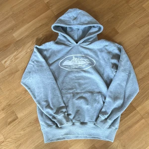 Grå hoodie från corteiz med tryck, medium - Grå hoodie i mjuk bomullsblandning med stor huva och magficka. På bröstet finns ett vitt tryck av en byggnad i oval ram. Klassisk passform och ribbade muddar vid ärmar och midja. Perfekt för chill dagar eller när du vill ha en clean streetstyle.