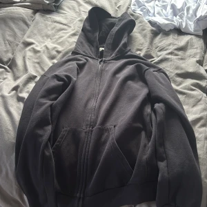 Svart hoodie med dragkedja - En klassisk svart hoodie med dragkedja framtill och huva. Tröjan har två stora fickor på magen och ribbade muddar vid ärmslut och nederkant. Perfekt för chill dagar eller när du vill ha något bekvämt och enkelt. Passar dig runt 170