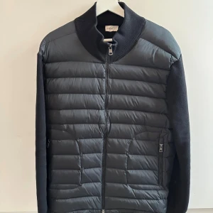 Svart pufferjacka från Moncler - Svart pufferjacka från Moncler med quiltad framsida och stickade ärmar och rygg. Jackan har hög krage, dragkedja framtill och två fickor med dragkedja. Snygg och stilren design med diskret Moncler-logga på ärmen.
