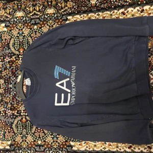 Blå EA7 Emporio Armani sweatshirt - Snygg marinblå sweatshirt från EA7 Emporio Armani med stor logga framtill i vitt och blått. Tröjan har rund halsringning och långa ärmar, perfekt för en chill och stilren look.
