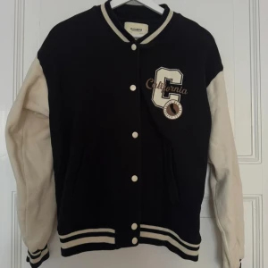 Svart och vit jacka från Pull&Bear - Collegeinspirerad svart och vit jacka från Pull&Bear med broderad 'California'-patch på bröstet. Jackan har kontrastfärgade ärmar, ribbade muddar och knäppning med vita knappar. Perfekt för en sportig och trendig look.