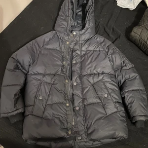 Svart pufferjacka från Zara Kids - Svart pufferjacka från Zara Kids med huva och stora fickor framtill. Jackan har både dragkedja och tryckknappar, samt quiltad design. Perfekt för kalla dagar och har en loose passform som ger en chill vibe.